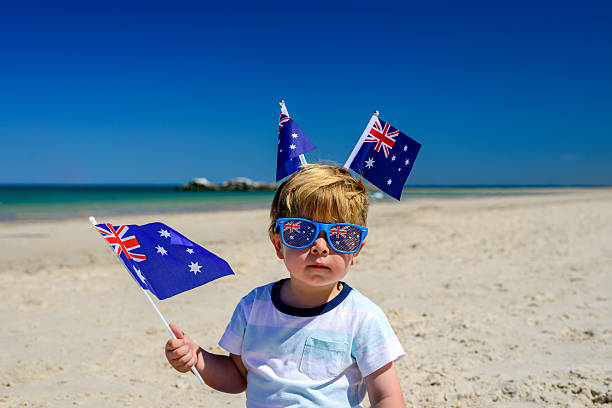 Cute smiling kid with Australian flags sitting on the sand at the beach on Australia Day