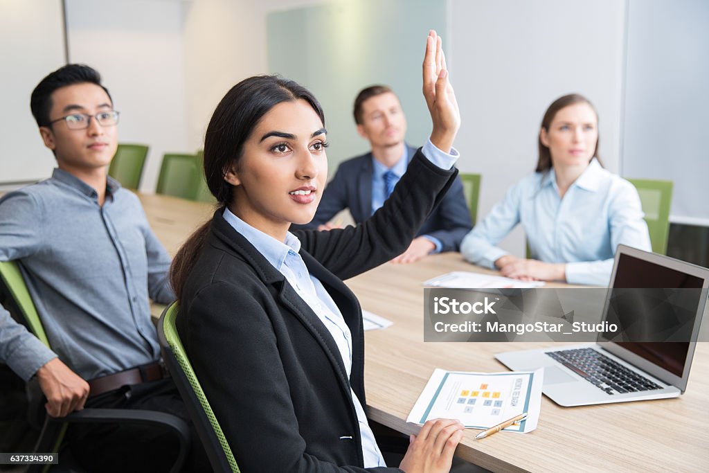 Confident young manager raising hand at workshop Confident young Latin-American female manager sitting at wjrkshop with multiethnic business people and raising hand to ask question Indian Ethnicity Stock Photo