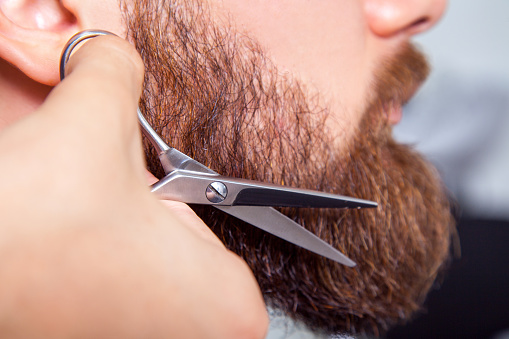 Barber with scissors shaving bearded man