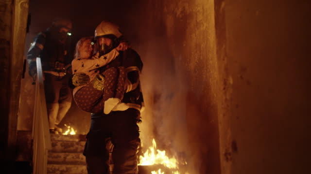 Burning Building. Group Of Firemen Descend on Burning Stairs. One Fireman Holds Saved Girl in His Arms.
