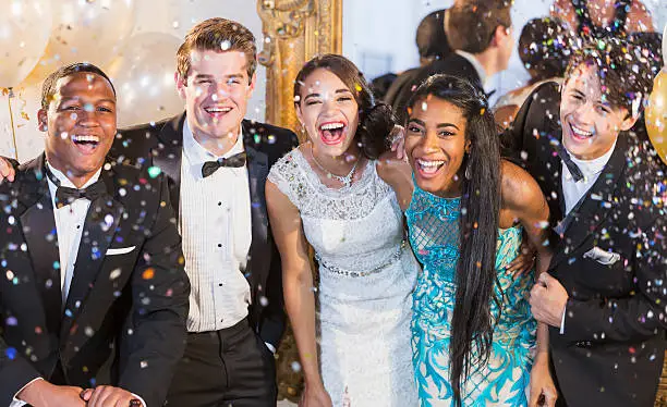A group of five multi-ethnic teenagers and  young adults dressed in formalwear - dresses and tuxedos. They are at a special event, a party or prom, standing together in a row, laughing and smiling. Or maybe a New Year's Eve party, confetti in the air to celebrate.