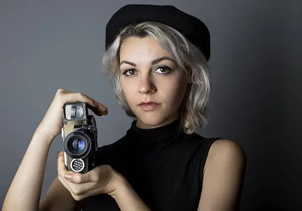 Woman holding a vintage video camera posing as a director, filmmaker, or cinematographer in the hollywood movie industry.  The image depicts creative arts.  She is wearing a hat and dressed like a noir artist while holding a retro classic lens.