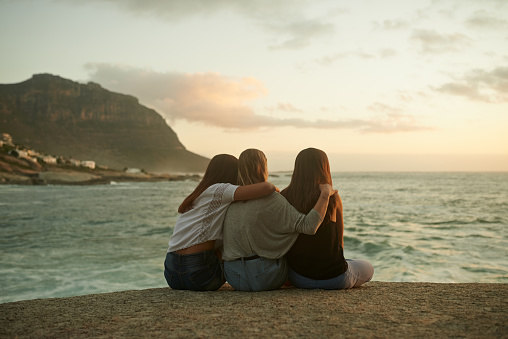 Shot of three friends admiring the view while sitting on a rock by the ocean