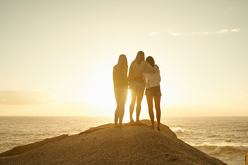 Rearview shot of three friends admiring the view while standing on a rock by the ocean