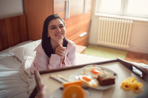 Beautiful young woman in pajamas sitting in her bed and smiling, surprised with breakfast in bed
