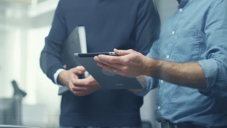 Two Senior Engineers Discussing Technical Details over Tablet Computer. Both Men Look Respectable and Professional. Office is Bright and Modern.