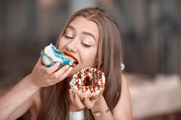 eating delicious donuts cute young woman holding donuts and biting it.cravinf for sweets.horizontal lifestyle shot. hungry stock pictures, royalty-free photos & images
