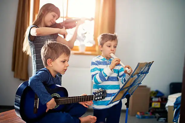 Sister and two brothers having fun playing music together. Sun is shining through the window. The girl is aged 10 and the boys are aged 6. Kids are playing a guitar, a violin and a flute.