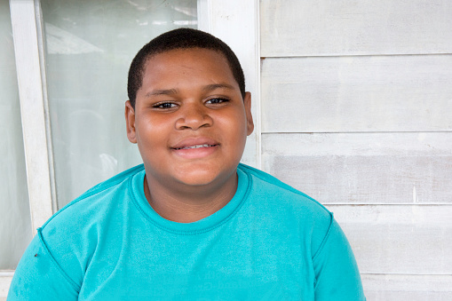 Eleven year old boy from the Honduran island of Roatan is sitting outside in front of a window and white wall posing for the camera. He is overweight. Taken with Canon 5D Mark lll.