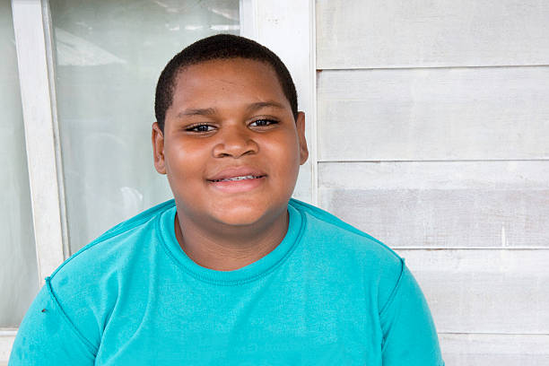 Series:Pre-adolescent Honduran boy with large build Eleven year old boy from the Honduran island of Roatan is sitting outside in front of a window and white wall posing for the camera. He is overweight. Taken with Canon 5D Mark lll. overweight boy stock pictures, royalty-free photos & images
