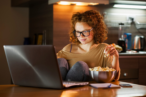 Young red hair woman working on computer late at night and eating popcorn