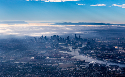 Melbourne skyline with skyscrapers emerging from the morning fog. Early morning aerial view overlooking Yarra river, harbor area and city center. Yarra Ranges National Park is covered by low clouds in the background.