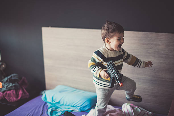 Fun with my toys! Baby boy playing with a gun and jumping on the bed baby gun stock pictures, royalty-free photos & images