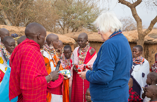 Selenkay, Kenya - October 9, 2016: White haired European female tourist with man, women and children in Maasai village. The women are dressed in traditional colorful kangas and pearl jewelry.  She is standing talking to one of the senior women, looking at a pearl necklace that she has been given. Cow's dung huts in the background.