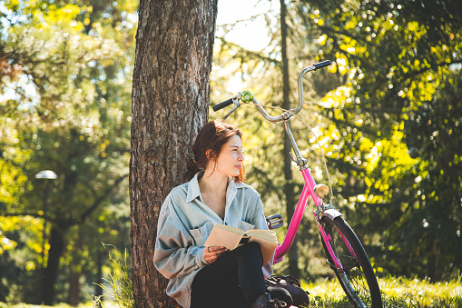 Beautiful young girl sitting in the forest, reading a book and enjoying the sunny spring day outside of the city.