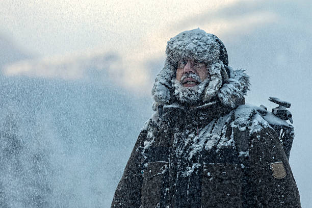 Man with furry  in snowstorm with cloudy skies and snowflakes Man with  furry and coat shivering in storm with cloudy skies and snowflakes blowing in wind polar climate stock pictures, royalty-free photos & images