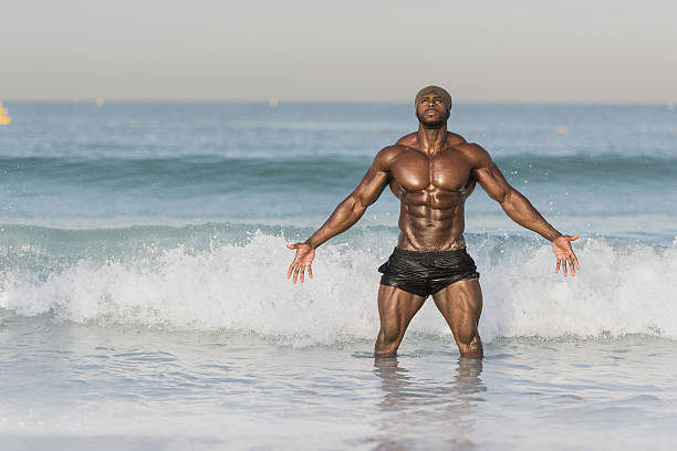 A dark muscular Middle eastern man, black man with oil on his body wearing a hair wrap being splashed by a wave, standing with hands out