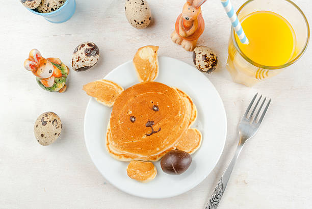 Pancakes for Easter breakfast Cute and fun for children at Easter Breakfast: Pancake made in the shape of a bunny with chocolate egg in the legs. On a white table, with a glass of juice, surrounded by toys & decorations. Top view bunny pancake stock pictures, royalty-free photos & images