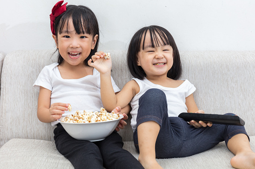 Happy Asian Chinese little sisters eating popcorn on the sofa in the living room at home.