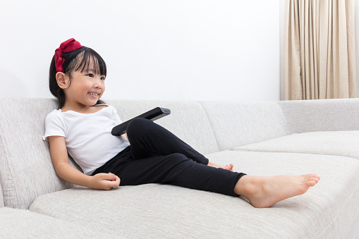 Happy  Asian Chinese little girl holding a TV remote control sitting on the sofa in the living room at home.