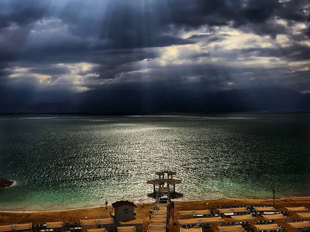 Dead Sea deserted beach. Rays of sunshine breaking through storm clouds at the Dead Sea – bordering Israel, the West Bank and Jordan – is a salt lake whose banks are more than 400m below sea level, the lowest point on dry land. Its famously hypersaline water makes floating easy, and its mineral-rich black mud is used for therapeutic and cosmetic treatments at area resorts. The surrounding desert offers many oases and historic sites.
