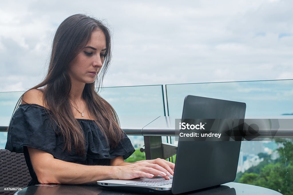 Young female freelancer dressed in black working on a project Young female freelancer dressed in black working on a project on laptop computer while sitting at rooftop cafe terrace. Adult Stock Photo