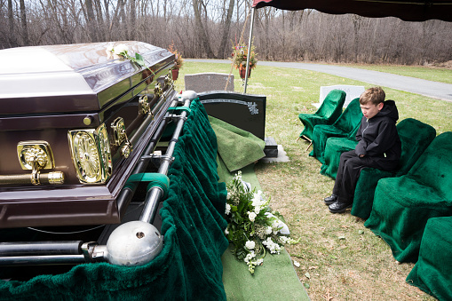 Small Caucasian boy 8-10 years with a sad expression sits alone in a row of fabric covered chairs under an awning and stares at a casket prepared for burial after a funeral ceremony in a cemetery in the Midwest on a cold spring day, USA
