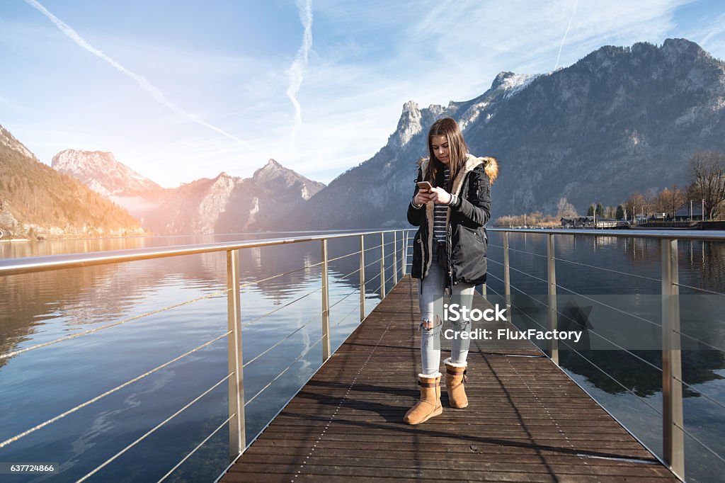 Young girl using smartphone in nature near lake Traunsee lake in Austria , in background are Traunstein mountains , 16 year old girl using phone on a dock in lake , posting photo on internet Girls Stock Photo