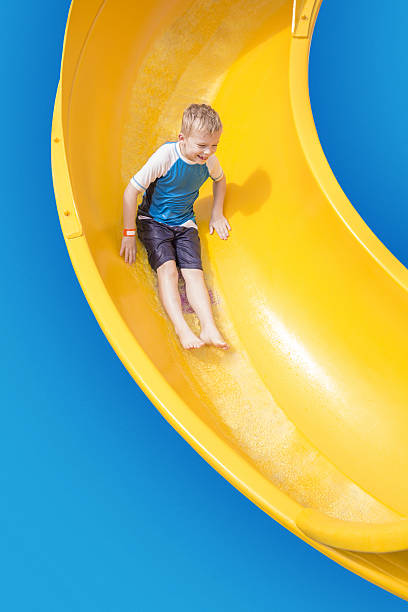 Smiling Young boy riding down a yellow water slide A cute young boy riding down a yellow water slide at an outdoor waterpark. Lots of copy space and fun colors sliding down stock pictures, royalty-free photos & images