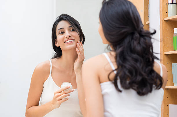 Woman applying moisturizer Woman caring of her beautiful skin on the face standing near mirror in the bathroom. Beautiful young woman applying moisturizer on her face. Smiling girl holding little jar of skin cream and applying lotion on face. exfoliation photos stock pictures, royalty-free photos & images