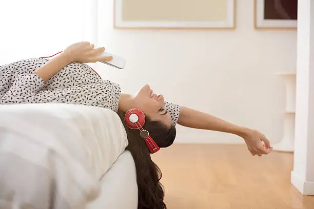 Happy young woman with headphones listening to music while lying on bed at home. Beautiful brunette girl with headphones relaxing on the bed. Smiling woman enjoying music on headphones holding phone and stretching out arms in bedroom.