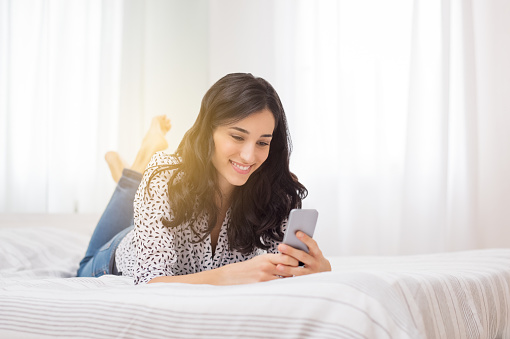 Smiling woman looking at mobile phone lying down on white bed. Happy brunette young woman using cellphone at home. Beautiful girl typing on smartphone and surfing the net.