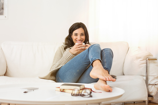 Happy woman sitting on couch holding a cup of tea in living room. Portrait of a young woman drinking a cup of coffee while relaxing on sofa at home. Smiling girl with woolen jacket drinking hot coffee and looking at camera.