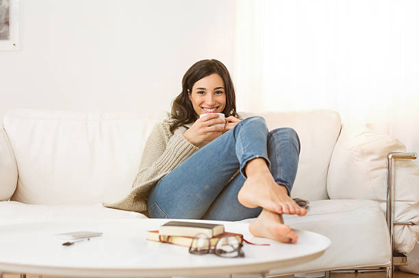 Woman relaxing at home Happy woman sitting on couch holding a cup of tea in living room. Portrait of a young woman drinking a cup of coffee while relaxing on sofa at home. Smiling girl with woolen jacket drinking hot coffee and looking at camera. barefoot stock pictures, royalty-free photos & images