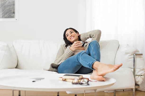 Smiling woman thinking on sofa Young smiling woman sitting on sofa and looking up while drinking hot tea. Young brunette woman thinking at home in a leisure time. Happy girl relaxing at home on a bright winter morning. Barefoot stock pictures, royalty-free photos & images