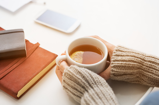 Close up of woman hands holding a cup of lemon tea on white table in a winter afternoon. Woman hands holding hot drink on table. Female hands with beige warm sweater holding hot tea with a slice of lemon.