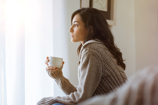 Young woman looking over window pane holding coffee. Thoughtful woman thinking and looking away while drinking hot tea. Woman in warm sweater looking outside window while drinking tea at morning.