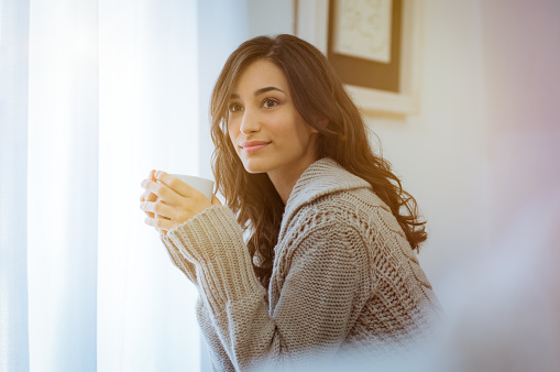 Smiling woman drinking hot tea while looking away. Happy young woman enjoying hot drink on a bright morning at home with warm sweater. Pensive girl sitting in front of the window while drinking hot coffee in a winter afternoon.