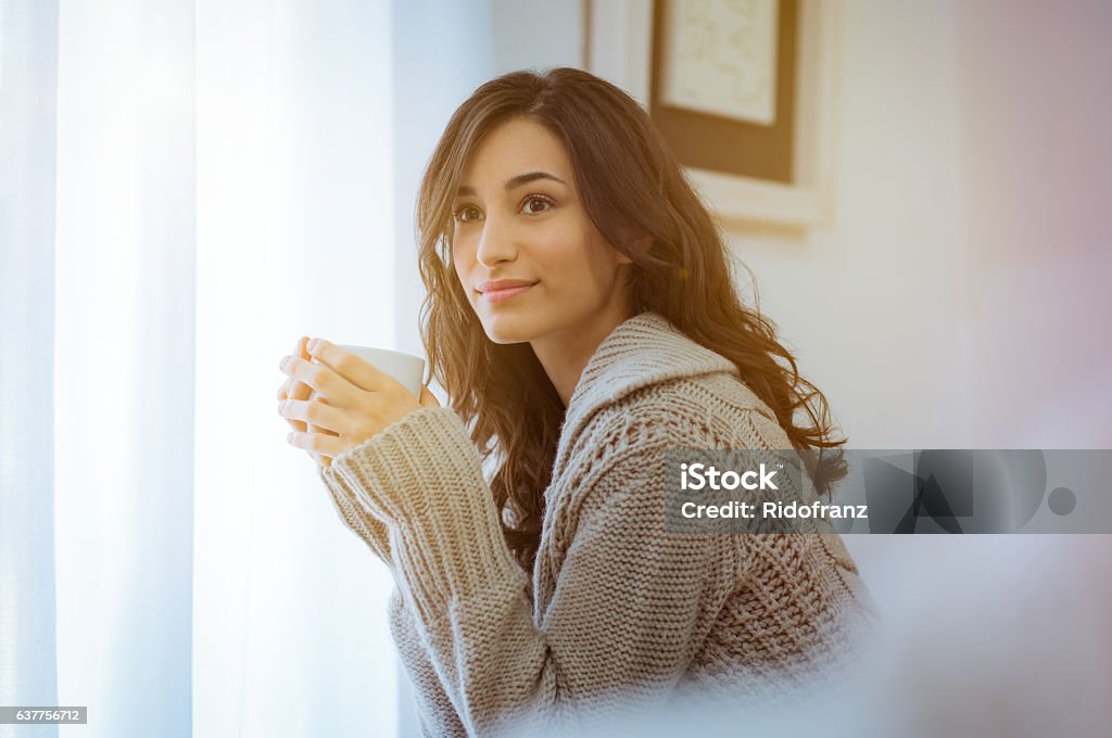 Woman enjoying morning coffee Smiling woman drinking hot tea while looking away. Happy young woman enjoying hot drink on a bright morning at home with warm sweater. Pensive girl sitting in front of the window while drinking hot coffee in a winter afternoon. Women Stock Photo