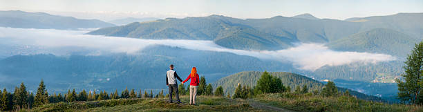 Back view man holding hands red-haired woman standing on a hill, enjoying a morning haze over the beautiful mighty mountains. Panorama