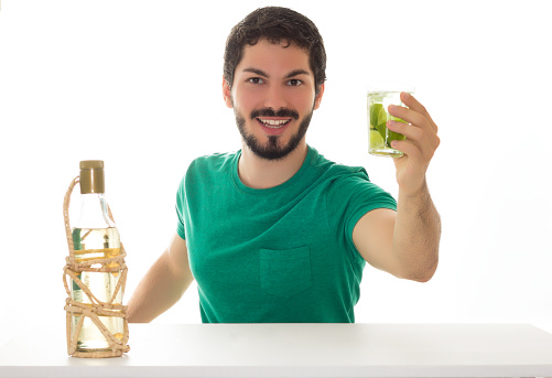 Young adult holding a cup of caipirinha and looking at the camera. Table and white background and a bottle of homemade cachaca