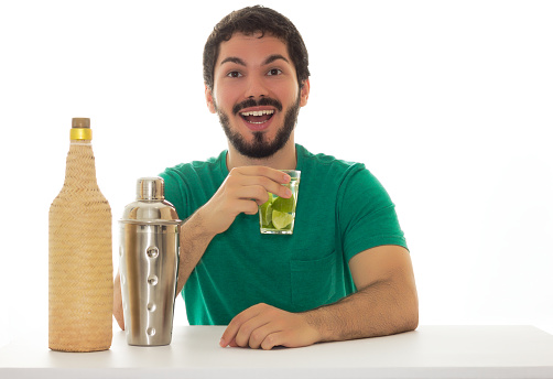 Young adult is holding a glass of drink made of lemon. Wearing green shirt. Table and white background; A bottle of homemade cachaca and a cocktail shaker.