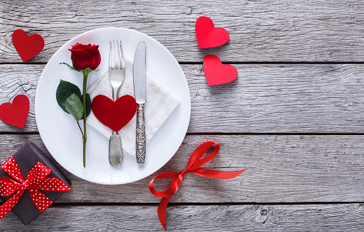 Romantic dinner concept. Valentine day or proposal background. Top view of restaurant wooden table with heart and rose with cutlery on plate. Copy space on rustic wood