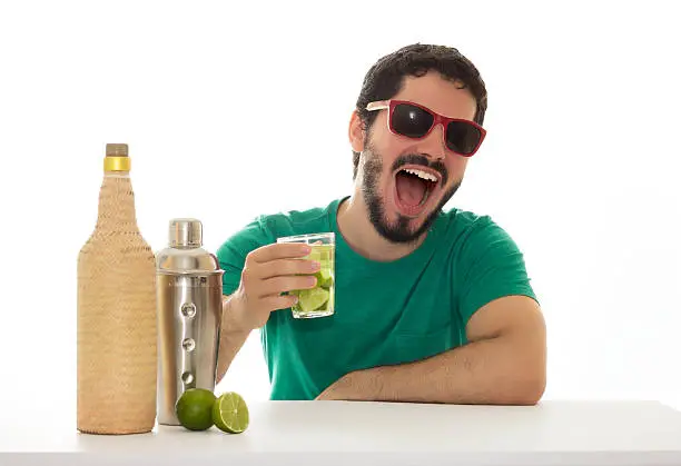 He is showing a glass with a drink made of lemon. Wearing green shirt. Table and white background; A bottle of homemade cachaca and a cocktail shaker.