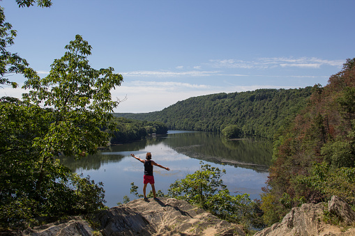 New Milford, United States- August 8, 2016. A man wearing a black tank and red shorts staring the landscape. View from a hiking trail in the east coast, with a lake, mountains, some rocks under a blue sky