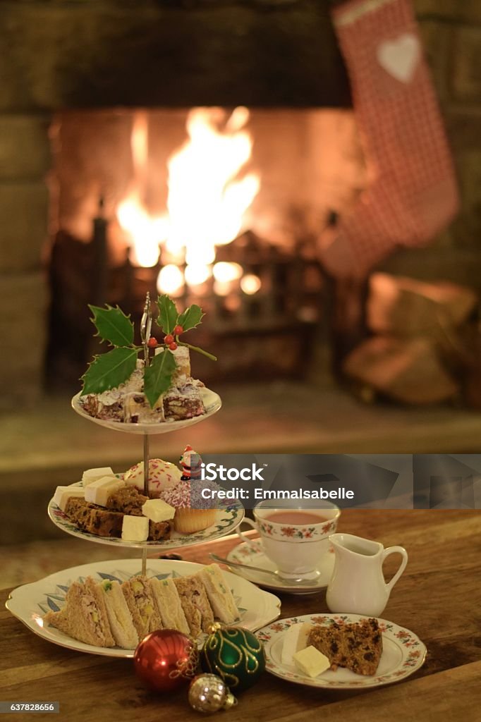 Festive Fireside Afternoon Tea Photograph of a table set for afternoon tea at Christmas time. There is a three tiered cake stand containing sandwiches, Christmas cake, miniature stollens and cup cakes. At the top is a sprig of holly. Also on the table is a cup of tea, a milk jug and a plate with Christmas cake and Wensleydale cheese. There is a small group of green, red and gold baubles beside the cake stand. In the background, an open fire can be seen with a stocking hanging beside it. Afternoon Tea Stock Photo