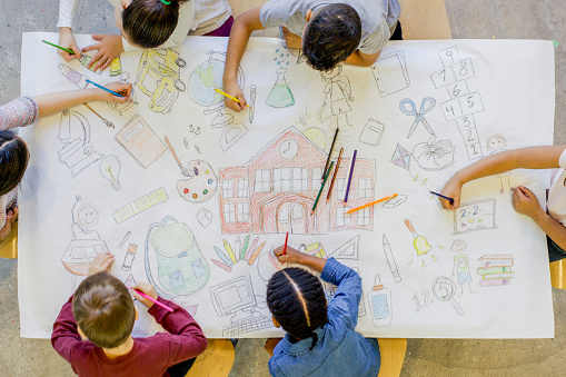 An aerial view of a multi ethnic group of children color in a series of back to school related images. The artwork includes pictures of a backpack, microscope, paints, bus, scissors, art supplies, and books.