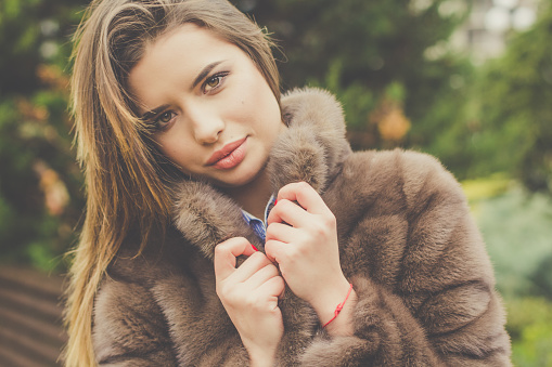 Portrait of beautiful teen girl with plump sexy lips is wearing warm brown winter fur coat posing in winter park. Fall and winter fashion concept.