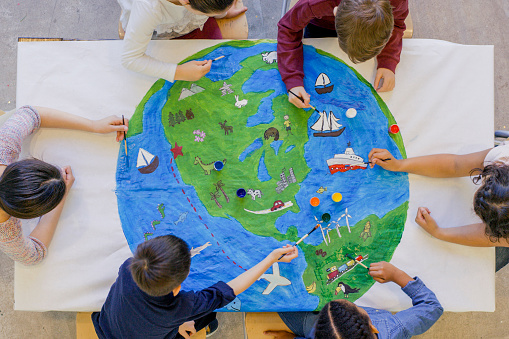 An aerial view of a multi ethnic group of children painting the globe and it's animals, fish, and vehicles. One girl paints a steam boat, while a boy colours in the ocean.