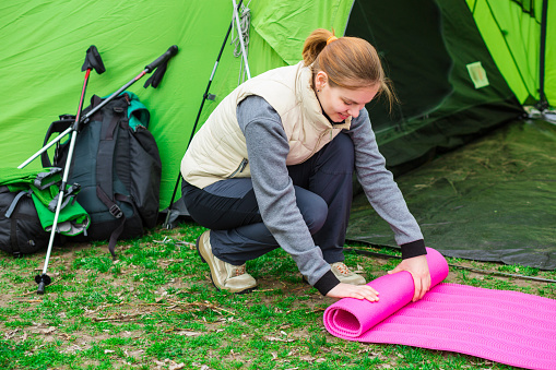 Cheerful woman rolling camping mat , during the camping holidays. In the background tourist tent and equipment for outdoor activities.